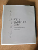 Ring binder titled 'A Year of Home Educating Our Way' on a cork surface