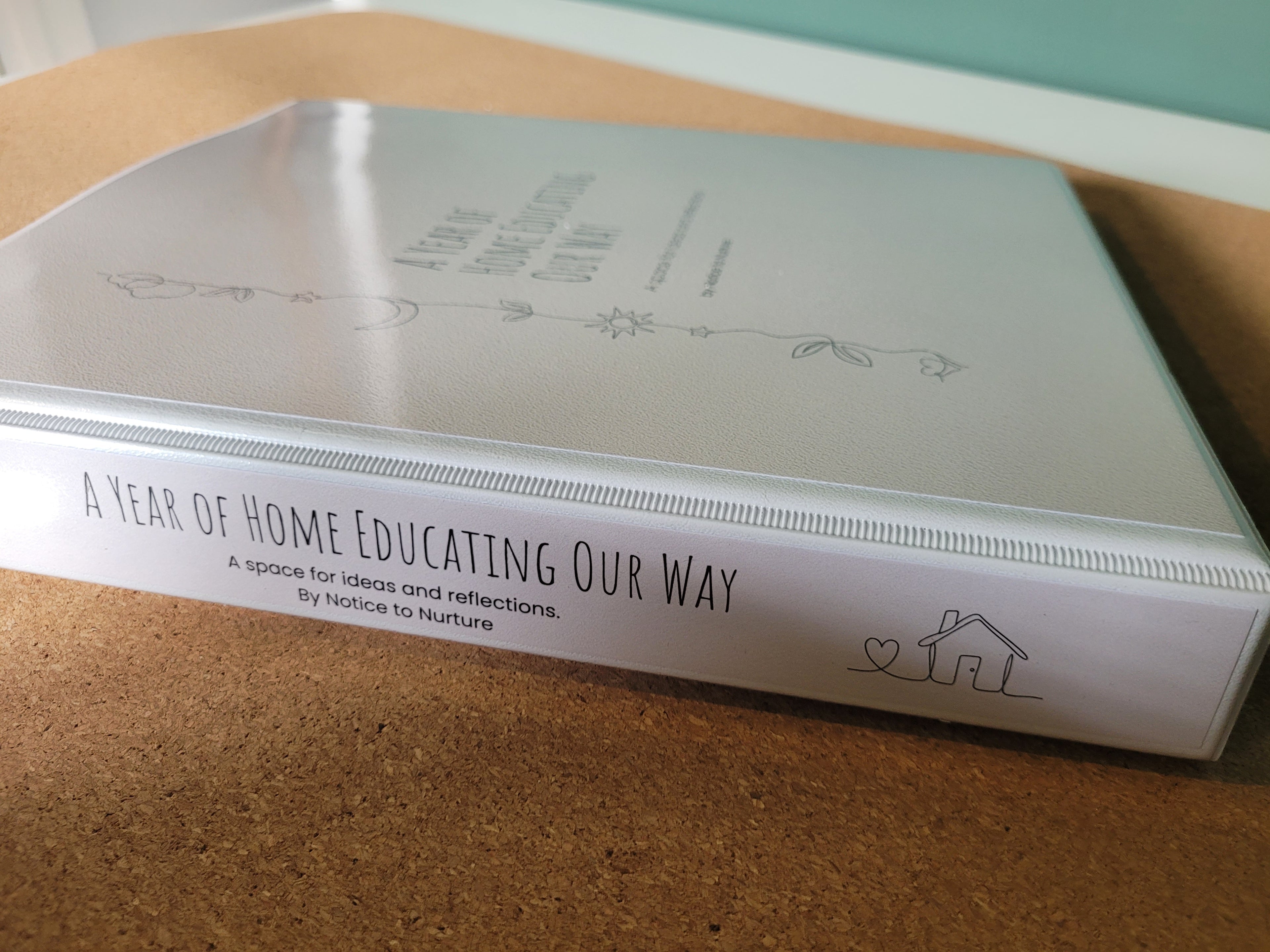 Ring binder titled 'A Year of Home Educating Our Way' on a cork surface