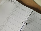 Open ring binder showing calendar page with a grid layout on a cork board background